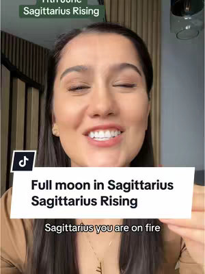 A woman with dark hair and a light brown top smiles. The background is blurred. A text overlay reads, "Full moon in Sagittarius Sagittarius Rising".