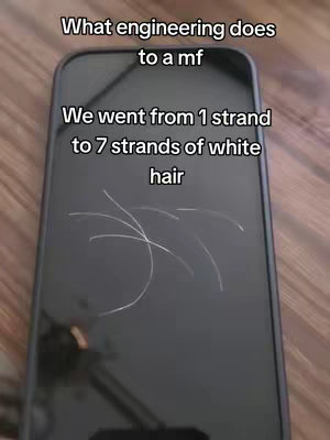 The image displays a close-up of a dark smartphone screen, appearing to be black. Overlaid on the screen are strands of white hair, suggesting the impact of engineering studies. Above, white text reads, "What engineering does to a mf" and "We went from 1 strand to 7 strands of white hair." The background is a wooden surface.