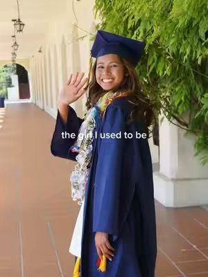 A young woman in a graduation gown and cap, standing outside with a bright smile and waving. She has a colorful lei around her neck. The background shows an outdoor walkway with columns and green foliage.