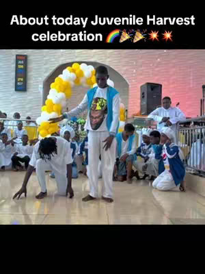 A photograph captures a church interior during what appears to be a celebration. A young man at the center, dressed in white pants and a white shirt with a printed design, gestures with his right hand, possibly addressing a crowd. He is surrounded by other individuals in white and blue uniforms, some kneeling, others standing. A decorative arch of yellow and white balloons frames the scene. The lighting is bright, and the background features church architecture.