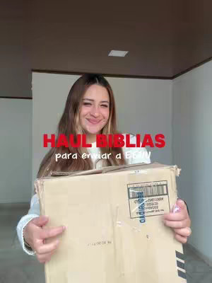 A young woman stands smiling, holding a large cardboard box in front of her. The box covers most of her body, but her face and shoulders are visible. She has long brown hair and is wearing a light-colored sweater. Above her head, overlaid in red text, is the hook 'HAUL BIBLIAS'. The background is a neutral-colored interior with a brown ceiling and white walls.