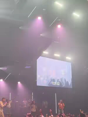 A low-angle shot of a church service. The image is dimly lit, with pink light and spotlights shining down. A large screen in the center displays text. People are visible in the foreground with their hands up.