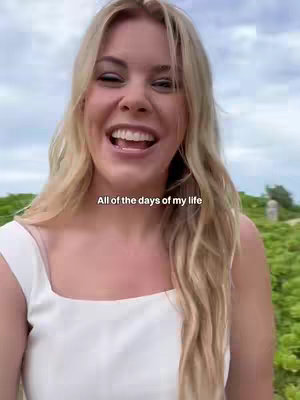 A woman with long blonde hair and a white dress is smiling widely. The background is blurred, suggesting an outdoor setting with greenery. Text overlayed near the top says, 'All of the days of my life'.