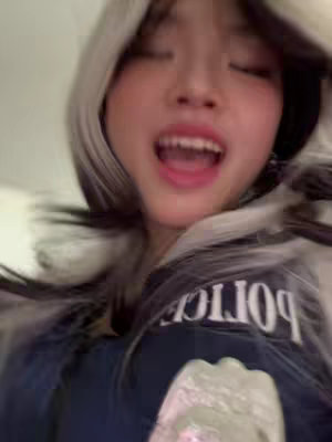 A young woman is the central focus, framed from the chest up. Her expression is animated, with her mouth wide open, suggesting she is singing or speaking enthusiastically. Her hair is styled with a two-tone effect, dark roots and white, framing her face. She is wearing a dark blue garment that has the word "POLICE" visible in white, and has a badge visible at the bottom of the frame.