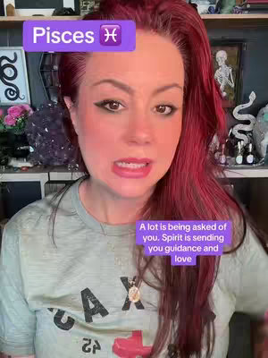 A woman with red hair is the focus of the selfie-style image. She has fair skin, bold makeup, and a slightly concerned expression. She is wearing a light gray t-shirt. Overlaid on the image are two text boxes: the first one features the word "Pisces" in white text inside a purple rectangle with the Pisces astrological symbol. The second, smaller box, appears at the bottom and reads 'A lot is being asked of you. Spirit is sending you guidance and love.' The background is a cluttered indoor space with shelves, decorative objects, and is softly lit.