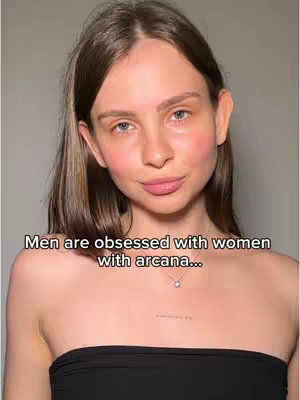 The cover image is a medium shot of a fair-skinned woman against a plain, light-colored background. She has long, light brown hair that is parted to the side. Her facial features are soft, with rosy cheeks and a neutral expression, and she is gazing directly at the viewer. She is wearing a strapless black top and a delicate necklace with a small pendant. Directly below her sternum is a small, unidentifiable tattoo. The text overlay, "Men are obsessed with women with arcana...", is placed at the bottom of the frame.