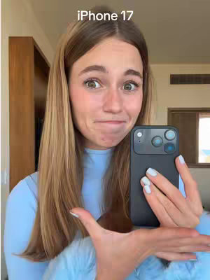 A young woman with long blonde hair is holding a black smartphone with three camera lenses. The woman is looking at the camera with a surprised expression. The background is a blurred room.
