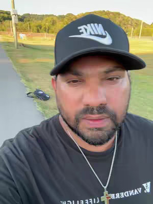A man wearing a black Nike cap and a black t-shirt, is standing on a paved path in an outdoor setting with green surroundings. He is looking down and slightly to his right. A pair of black shoes are lying beside a path. The man wears a necklace with a cross pendant.
