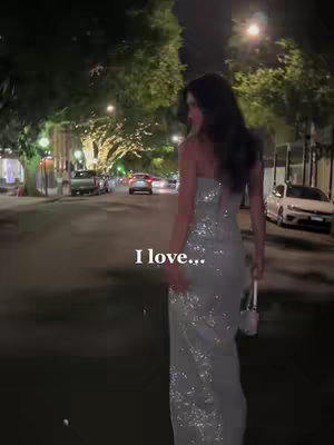 A woman in a sequined gown walks towards the camera down a city street at night. The road is lit by streetlights, and cars and buildings are in the background.