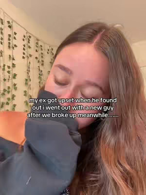 A close-up shot of a woman with long brown hair, looking down with her eyes nearly closed. She's wearing a dark hoodie, and her left hand is partially obscuring her face. The background is softly blurred, with hints of a wall decorated with lights and plant elements. Text overlayed at the bottom of the image states: "my ex got upset when he found out i went out with a new guy after we broke up meanwhile..."