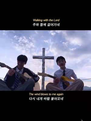 Two young men sit on an elevated surface, each playing an acoustic guitar. Behind them stands a simple wooden cross against a partially clouded sky. Lyrics are overlaid in Korean and English.