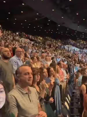 A live-action video from an event or gathering, with a blurred background of the audience and stage, a crowd of people are in the front.