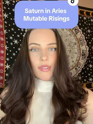 A woman with long brown hair and light eyes looks directly at the camera. She wears a white top and there is a star-patterned black background and a decorative tapestry behind her. A light blue text box at the top of the screen says "Saturn in Aries Mutable Risings".