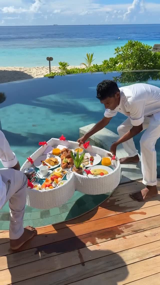 Two individuals are shown setting up floating trays of breakfast food in a pool. The trays are heart-shaped and filled with various breakfast items, including fruit, pastries, and orange juice. The scene is set at a luxury resort with an infinity pool overlooking a clear blue body of water, suggesting a tropical location.
