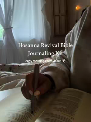 The image captures a cozy, intimate scene of someone engaged in Bible journaling. A person is seated in bed, illuminated by soft, warm lighting, possibly from a nearby lamp or ambient room light. The central focus is a hand delicately holding a pen over the open pages of the Bible. The composition utilizes a shallow depth of field, where the Bible and the hand are sharply in focus, while the background, including a white curtain and subtle details like a candleholder, gently fades into the background. The overall palette is warm and inviting, accentuated by the neutral tones of the bedding and the soft lighting casting shadows, creating a calming and focused atmosphere, ideal for reflection and study.