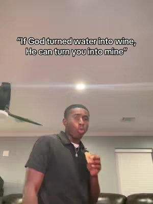 A man with short, neatly styled black hair, wearing a black polo shirt, stands in a domestic setting. He holds an unidentifiable object close to his mouth as if about to eat it. Above him, white text appears against a blurred view of the ceiling, providing a caption. The composition is a medium shot that captures the man from the chest up, with a ceiling fan visible in the background. The lighting seems to be from an overhead source, indicating a casually lit environment.