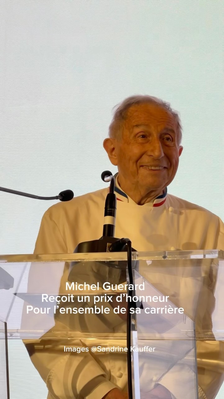 The cover image features a medium shot of an older man in a white chef's coat standing behind a clear podium. The man is smiling, and text overlays indicate he is receiving an honorary award. The background is a nondescript light color. Two microphones are visible near the man's face.