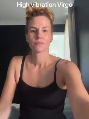 A woman with light brown hair styled in a bun is centered in the frame, wearing a black tank top. The background includes a window with light streaming in and a dark colored wall. Text overlayed at the top reads 'High vibration Virgo'.