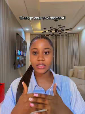 A woman with a ponytail and blue shirt is centered in a well-lit living room setting, with a text overlay that reads "Change your environment!" The woman is looking at the camera and using hand gestures while speaking, suggesting a direct and engaging presentation style; the background indicates an affluent living space with large TV.