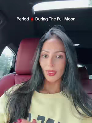 A woman with long dark hair is seated in a car, looking towards the camera. She has light skin and is wearing a yellow shirt. The car interior is visible, with red seats and a dark-colored dashboard. Above her is a sunroof. Text overlay at the top reads "Period 🩸 During The Full Moon".