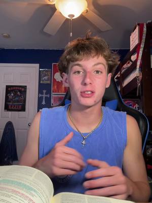A young man in a blue tank top sits indoors, speaking into a camera. He has short, light-brown hair and a gold chain with a cross pendant. In front of him is an open book. The background includes a ceiling fan, door, and wall art.