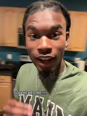 A man with a goatee is centered in the frame, looking directly at the camera with an open mouth and a slight smile. The background elements are out of focus, including a kitchen setting with a microwave and cabinets. A text overlay 'YOU HEARD ME!!!' is at the bottom, and he wears a green shirt and a silver chain.