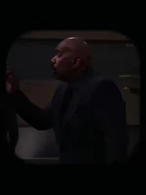 The image features a man, appearing to be of African descent and wearing a dark-colored suit or blazer, gazing upwards and slightly to the side. He is positioned in a setting that looks like an interior space, possibly an office or studio, suggested by the dimly lit background. The composition has a cinematic feel with the use of shadows.