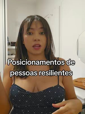 A woman with long brown hair is centered in the frame, looking towards the camera and speaking. Text overlayed on the image reads:

'Posicionamentos de pessoas resilientes'. The background is a room with a neutral color palette, suggesting an interior setting captured on a phone camera.