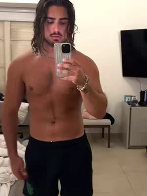 A shirtless man with long, wavy hair and a mustache stands in a room, taking a selfie with a smartphone held in his right hand. He wears black shorts and a silver chain and wristband. Behind him is a partially visible room with a window and a television.
