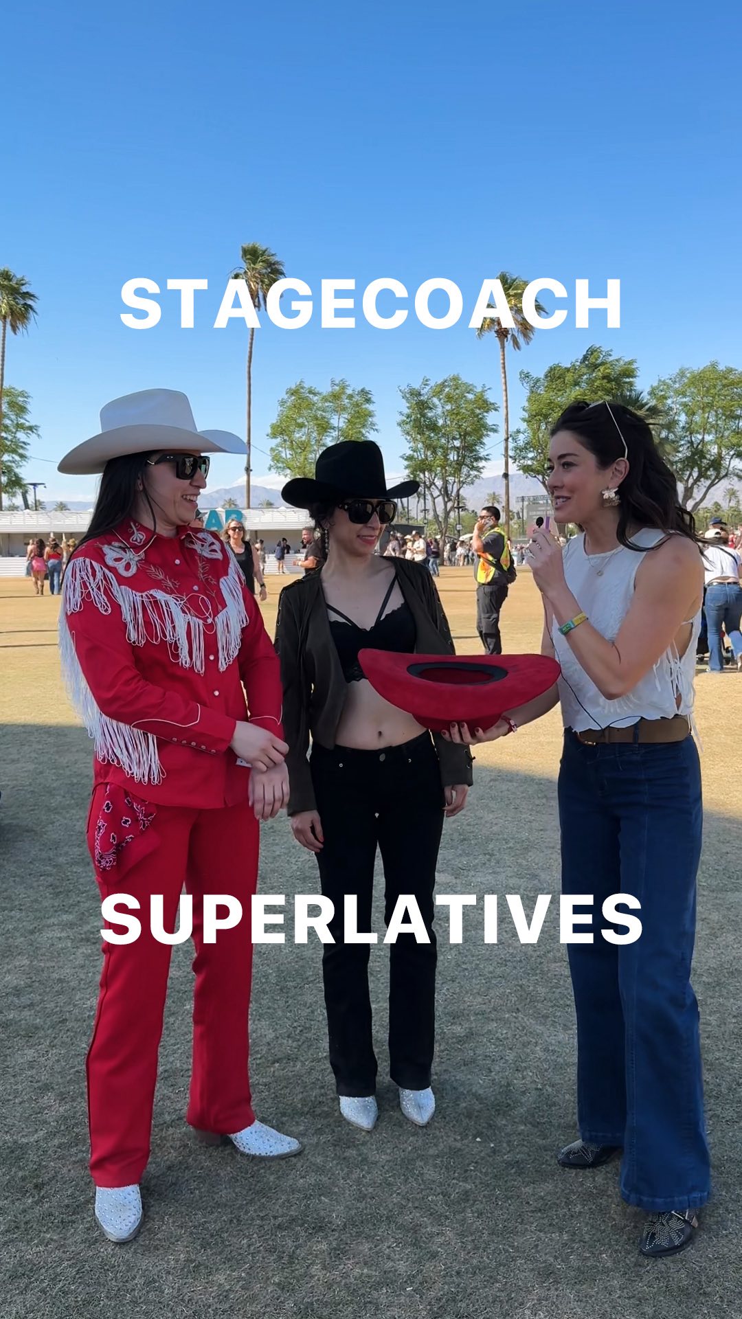 Three women are standing outside, presumably at a music festival, with trees and attendees in the background. The woman on the left is wearing a red outfit with white fringing on her jacket, a white cowboy hat, and sunglasses. The woman in the middle is wearing a black outfit with a black cowboy hat and holding a red hat. The woman on the right is wearing a white top and jeans, holding a microphone. Overlaid on the image are the words 'STAGECOACH' and 'SUPERLATIVES'.