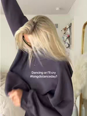 A woman with blonde hair and a dark sweatshirt is dancing. The background is a light-colored room with a mirror and various items.
