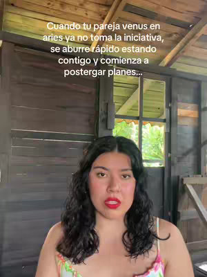 A woman stands indoors with a wooden background, possibly a shed or rustic structure. She has dark, curly hair, fair skin, and red lipstick. She wears a floral patterned top. Text in a yellow box highlights a relationship topic of zodiac signs.
