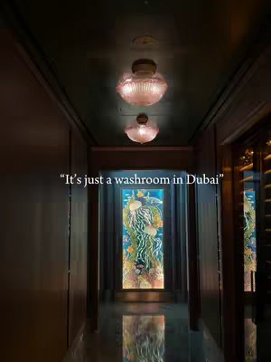 An interior shot down a hallway with a high gloss tile floor leading to a decorative glass panel illuminated from behind. The hallway is lined with dark wood paneling and doorways on either side. Two circular lights hang from the dark ceiling. Text is overlaid on the image in a small white font.