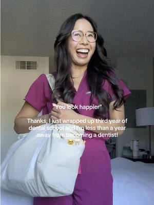 A young woman with long dark hair and glasses smiles with her mouth wide open, revealing teeth. She is wearing a magenta scrubs and holding a white bag. Text overlayed on image.