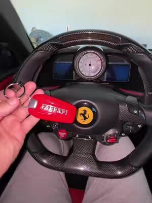 The cover image shows the interior of a car, focusing on the steering wheel and dashboard. A hand holding a red Ferrari key is in the foreground. The steering wheel is black and features a prominent Ferrari emblem. Various gauges and controls are visible, and the overall color scheme is dark with red accents.
