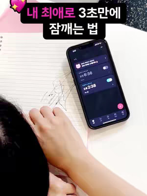 The image shows a person's arm and part of their face resting on a surface, possibly indicating they are tired or just waking up. Next to them, a smartphone displays an alarm application interface, implying content related to the app's function. The smartphone is held with the screen on, while also showing some scribbles on a notepad. The image has a warm, soft lighting.