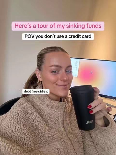 A woman with blonde hair in a ponytail is smiling at the camera as she holds a black travel mug. Text overlays read 'Here's a tour of my sinking funds' and 'POV you don't use a credit card'. The background is blurred, but suggests a home office setting.
