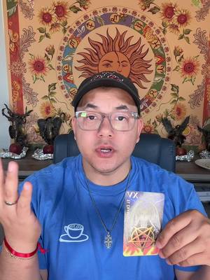 A man with glasses and a black cap is holding a tarot card, framed from the chest up. Behind him is a tapestry with a sun design. The man wears a blue T-shirt with a coffee cup design.