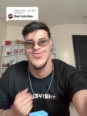 A selfie-style video cover featuring a young man with a distorted face filter. He's wearing sunglasses and a black t-shirt with white text. A comment bubble in the top left has the text "Deri tsta lina". The background shows a room with shelves and a door.