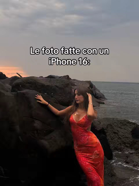 A woman in a red dress poses on rocks near the ocean at sunset. The sky is a mix of orange and grey, reflecting onto the water. The woman is positioned in the center, partially leaning on a dark rock. Text is overlaid at the top.