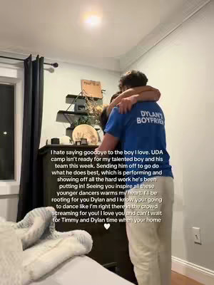 Two people in close embrace indoors. A person wearing a blue shirt with “DYLAN’S BOYFRIEND” printed on the back is hugging another person who is partially seen. The background includes a window with curtains, a dark-colored dresser, and a bed with a light-colored blanket. Text is overlaid, expressing affection and support for the individual named Dylan. The overall composition suggests a moment of affection and farewell.