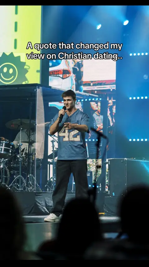 A man on a stage is speaking into a microphone, wearing a blue jersey shirt with the number 42 on it and black pants. Behind him, on a large screen, are blurred colors and shapes. The scene is lit with stage lighting.