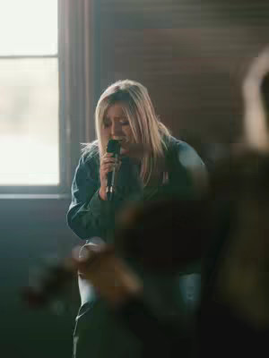 A woman with long blonde hair sings into a microphone, positioned center frame. She is wearing a long-sleeved shirt and looks downward while holding the microphone. Behind her is a window, and other blurred figures are in the background. The lighting is soft, with natural light. The mood is devotional.