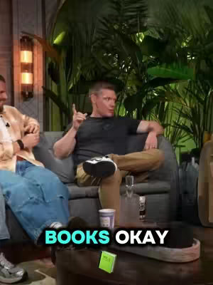 A medium shot of a man seated on a couch in an indoor setting, with another person partially visible to his left. The main subject is wearing a dark shirt and tan pants, with his left arm raised and index finger extended as if making a point. The lower portion of his legs and feet are visible, revealing sneakers. In the background, there's lush green foliage, a decorative lamp, and a small table with drinks and a book, suggesting a talk-show or interview setting. The text overlayed at the bottom of the image states "BOOKS OKAY" in a bold, white font.