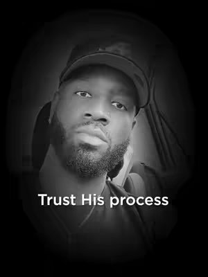 A grayscale portrait of a Black man in a car, looking directly at the camera with a serious expression. The man is wearing a baseball cap and has a neatly groomed beard. Text overlayed on the image reads "Trust His process".