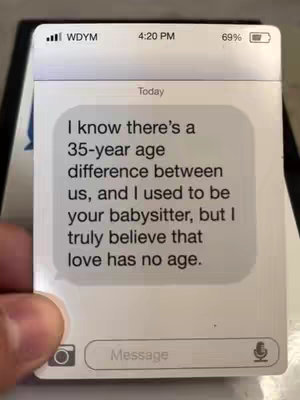The image shows a close-up of a phone screen displaying a text message conversation within a white card. The screen emulates an iPhone interface, with rounded corners and a status bar at the top showing the time "4:20 PM", the carrier name "WDYM", battery percentage, and Wi-Fi signal strength alongside a battery icon. The body of the text reads "I know there's a 35-year age difference between us, and I used to be your babysitter, but I truly believe that love has no age." The message is contained within a white text bubble over a plain light gray background. An image of a hand holding the phoneβs surface is visible on the bottom left. The colors are predominantly white and light gray, with small accents of black.