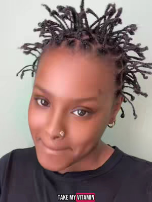 A close-up shot of a person with dark skin and face, wearing a black shirt, their hair styled into small, tightly-wound braids. They have a nose ring, and their expression is a slight, almost shy smile. Below the person’s face is a pink box with the text "TAKE MY VITAMIN". The background is a muted teal or green.