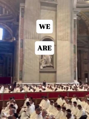 Interior shot of a large cathedral filled with rows of people in white robes. Above the crowd are two white rectangular boxes with the words 'WE' and 'ARE' in black. A tall stone pillar is on the left and a statue is on the right inside the church