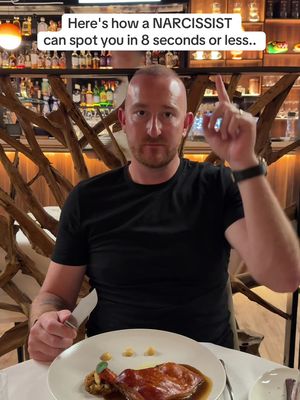 A man in a black shirt points upwards with his right index finger while looking at the camera. He is seated at a table in a restaurant, with a plate of food in front of him. The background consists of a bar with bottles and wooden decorations.