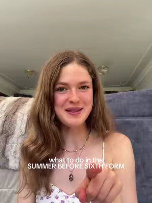 A young woman with long, wavy brown hair is centered in the frame, smiling and pointing towards the viewer. She wears a white tank top with a floral pattern and a necklace. The background includes a blurred white wall a neutral colored sofa and the ceiling with lights.